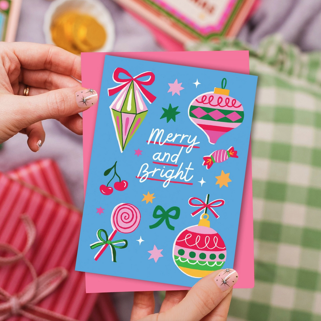 Hand holding a colorful greeting card with festive illustrations and 'Merry and Bright' text.